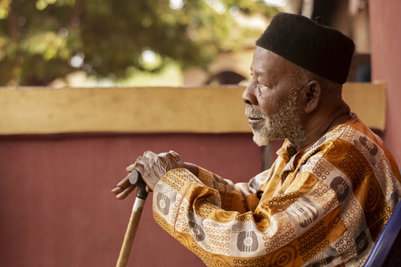 An older man seated outdoors, resting on a walking stick